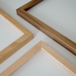 Op de afbeelding: Close-up van drie houten fotolijsten in verschillende bruintinten. De frames zijn schuin geplaatst, waardoor hun hoeken en de natuurlijke houtnerf zichtbaar zijn. Het licht reflecteert op de gladde oppervlakken, wat het vakmanschap benadrukt.