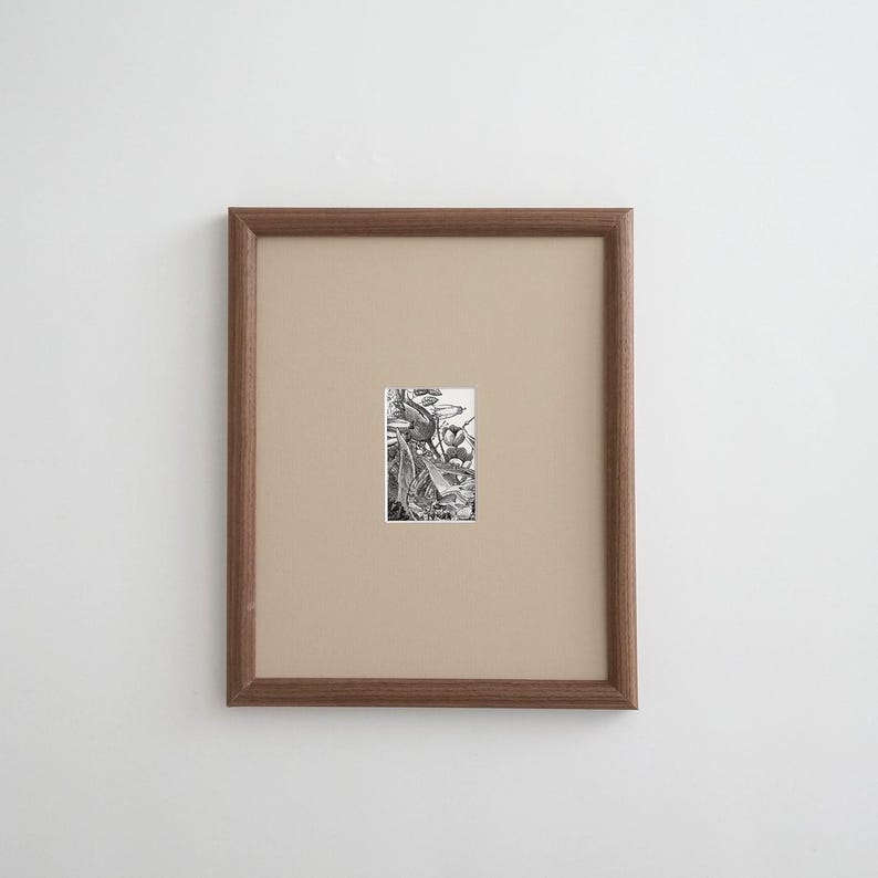K&ouml;nnte beinhalten: Ein rechteckiger Holzrahmen mit einem beigefarbenen Passepartout, das eine kleine, zentrierte botanische Illustration in Schwarzwei&szlig; umschlie&szlig;t. Das Kunstwerk zeigt detaillierte Strichzeichnungen verschiedener Pflanzen, Bl&auml;tter und Blumen.
