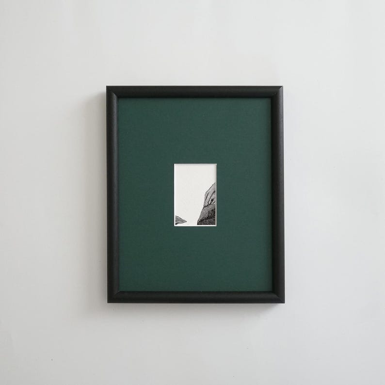 May include: A framed art piece featuring a black frame, a dark green mat, and a small black and white image. The image appears to be a section of a building or structure. The frame is rectangular and mounted on a white wall.