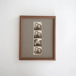 Op de afbeelding: Een rechthoekige houten fotolijst met een verticale strook van vier zwart-wit fotohokje-portretten. De strook toont een koppel in verschillende poses, waaronder glimlachend en kussend, geplaatst tegen een effen grijze passe-partout in de lijst.