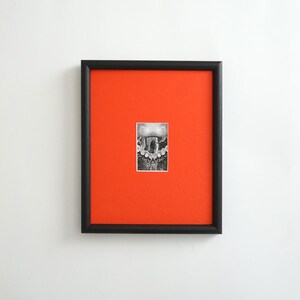May include: A framed black and white artwork with a small, detailed drawing centered on a vibrant orange background. The artwork is enclosed in a black frame, creating a striking contrast. The drawing appears to be a detailed illustration.
