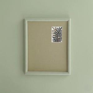 May include: A framed artwork with a light green frame. The artwork has a beige background and a small rectangular print of a botanical illustration in green, black, and gold. The frame is a pale green colour.