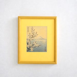 Op de afbeelding: Een ingelijste prent van een Japans landschap met de berg Fuji en kersenbloesems. Het kunstwerk zit in een gele lijst en toont een serene scène met een berg, water en bloeiende takken. De prent heeft Japanse tekst.