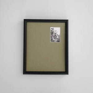 May include: A framed artwork with a black frame and a light olive-green mat. The artwork features a small, detailed black and white illustration. The frame is rectangular and mounted on a white wall.