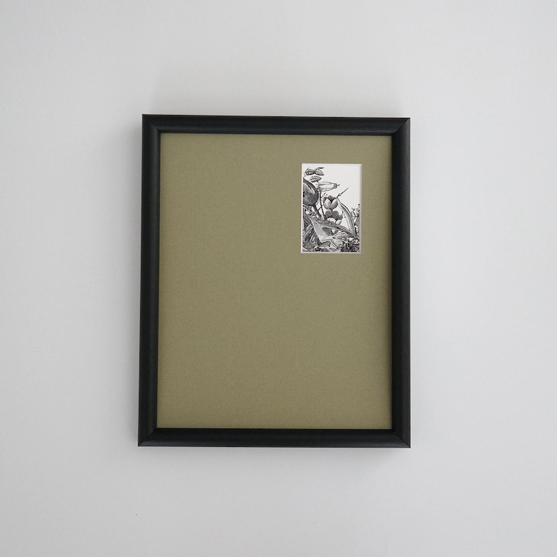 May include: A framed artwork with a black frame and a light olive-green mat. The artwork features a small, detailed black and white illustration. The frame is rectangular and mounted on a white wall.