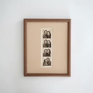 May include: A rectangular wooden picture frame with a tan mat board containing a vertical strip of four black and white photo booth images. Each frame in the strip shows a man and a woman posing together in various expressions and positions.