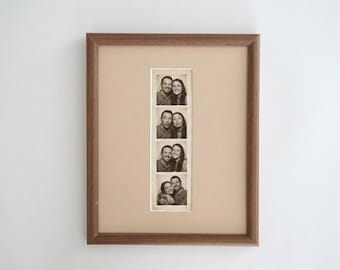 May include: A rectangular wooden picture frame with a tan mat board containing a vertical strip of four black and white photo booth images. Each frame in the strip shows a man and a woman posing together in various expressions and positions.