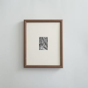 May include: A framed black and white print. The print is a detailed illustration with various figures and objects. The frame is a medium brown color, and the print is set against a cream-colored mat. The artwork is displayed on a white wall.