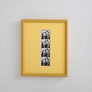 Könnte beinhalten: Ein rechteckiger Bilderrahmen mit matter gelber Oberfläche und passendem gelbem Passepartout. Zentriert im Rahmen befindet sich ein vertikaler Schwarz-Weiß-Fotostreifen mit vier einzelnen Bildern, die zwei Personen mit verschiedenen Gesichtsausdrücken zeigen.