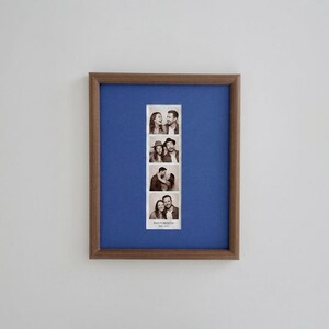 K&ouml;nnte beinhalten: Ein rechteckiger Bilderrahmen aus Holz mit einem blauen Passepartout, das einen vertikalen schwarz-wei&szlig;en Fotoautomatenstreifen mit vier Bildern von zwei Personen enth&auml;lt. Unten auf dem Fotostreifen steht der Text PHOTOBOOTH MAY 2023.