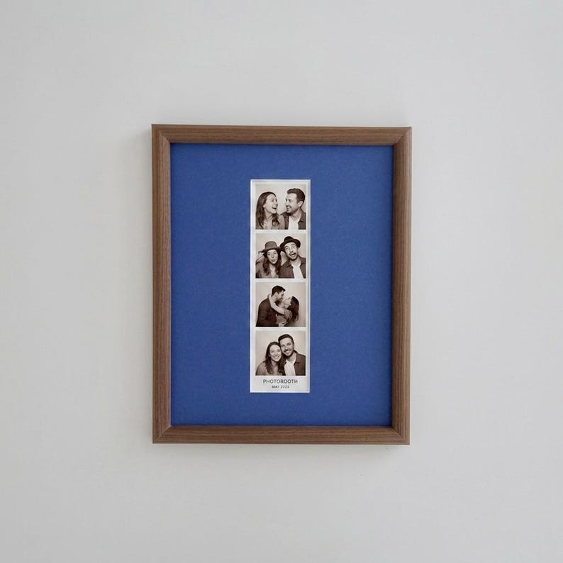 K&ouml;nnte beinhalten: Ein rechteckiger Bilderrahmen aus Holz mit einem blauen Passepartout, das einen vertikalen schwarz-wei&szlig;en Fotoautomatenstreifen mit vier Bildern von zwei Personen enth&auml;lt. Unten auf dem Fotostreifen steht der Text PHOTOBOOTH MAY 2023.
