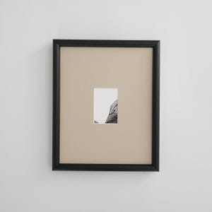 May include: A framed artwork featuring a small, rectangular black and white image. The artwork is set against a beige background within a black frame. The image appears to be a detailed drawing or print.