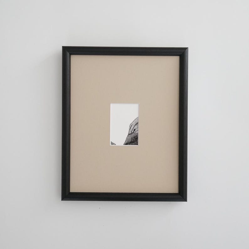 May include: A framed artwork featuring a small, rectangular black and white image. The artwork is set against a beige background within a black frame. The image appears to be a detailed drawing or print.