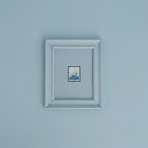 May include: A light blue rectangular picture frame containing a small postage stamp centered on a matching light blue mat. The stamp features a detailed blue and white illustration of a sailing ship with the text NORGE 50 ORE printed at the bottom.