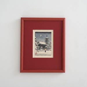 May include: Framed print of a Japanese winter scene. The artwork depicts a pagoda, snow-covered trees, and a person with an umbrella. The frame is a deep red color, and the print is matted in white.