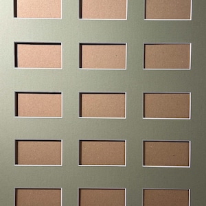 Può includere: Un passe-partout color verde salvia con una griglia di quindici aperture rettangolari disposte in cinque file e tre colonne. Ogni apertura è incorniciata da un sottile bordo bianco, che rivela il cartone marrone dietro le aperture.