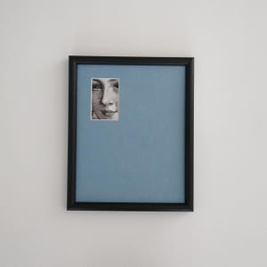 May include: A framed artwork featuring a portrait of a person's face. The portrait is a small, rectangular image set against a light blue background within a black frame. The frame is rectangular and mounted on a white wall.