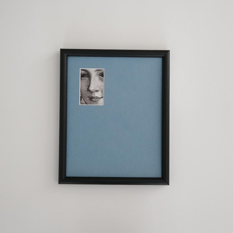 May include: A framed artwork featuring a portrait of a person's face. The portrait is a small, rectangular image set against a light blue background within a black frame. The frame is rectangular and mounted on a white wall.