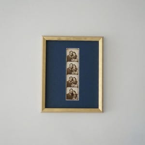 Op de afbeelding: Een gouden fotolijst met een zwart-wit fotostrip van een stel. De passe-partout is donkerblauw. De rechthoekige lijst hangt aan een witte muur.