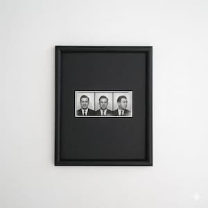 Op de afbeelding: Zwart omlijst kunstwerk met drie zwart-wit portretten van mannen in pakken. De foto's zijn horizontaal gerangschikt in een witte rand. De lijst is effen zwart.