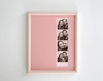 May include: A square picture frame with a light pink border and a matching pink mat board. The frame contains a vertical strip of four black and white photo booth images featuring a couple posing together in various expressions.