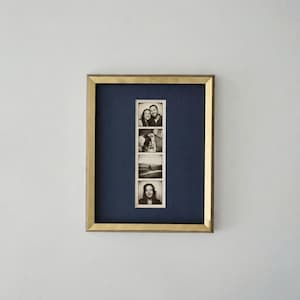 Op de afbeelding: Een gouden fotolijst met vier zwart-wit foto's in een strook. De lijst heeft een marineblauwe passe-partout. De foto's tonen verschillende mensen en een hond.