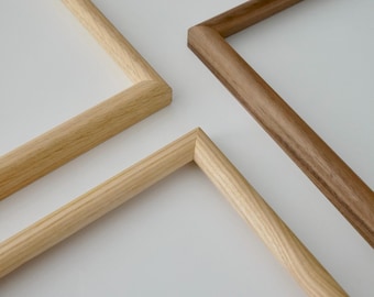 May include: Three wooden picture frames arranged in a corner-to-corner layout. Two frames are made of light-colored wood with a visible grain, while the third frame is made of a darker, rich brown wood with a smooth, consistent finish.