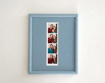 May include: A light blue rectangular picture frame featuring a vertical photo strip with four distinct frames. Each frame displays a series of candid portraits of two adults and a child, captured in various expressions against a white background.