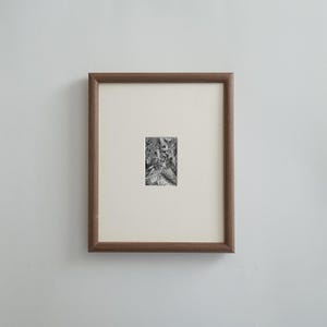 May include: A framed black and white artwork. The artwork is a small, detailed illustration set within a cream-colored mat. The frame is a dark brown wood. The artwork is centered within the frame and mounted on a white wall.