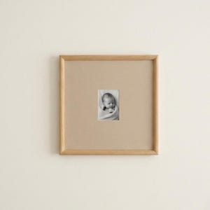 May include: A square light wood picture frame featuring a wide beige mat board. Centered within the mat is a small, vertical black and white photograph of a swaddled infant with eyes closed, resting with hands near the face.