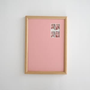 Könnte beinhalten: Ein rechteckiger Bilderrahmen mit einem hellen Holzrahmen und einem einfarbigen rosa Passepartout. In der oberen rechten Ecke des Passepartouts befindet sich eine kleine quadratische Collage mit vier einzelnen Fotos eines Babygesichts.