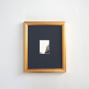 Peut inclure: Un cadre mural rectangulaire avec une finition dorée métallique et un passe-partout bleu marine foncé. Le cadre contient une petite impression blanche centrée représentant un dessin à l'encre noire et blanche d'une falaise rocheuse.