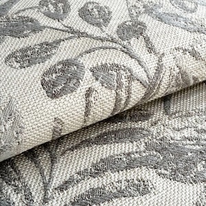 May include: Close-up of a woven fabric with a floral pattern. The design features gray leaves and flowers on a light beige background. The fabric is folded over, revealing its texture and construction.