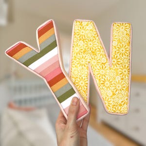 May include: Two fabric letters, "Y" and "N", are shown. The "Y" has a striped design in orange, pink, green, and grey. The "N" is patterned with yellow flowers. Both letters have a pale pink border.