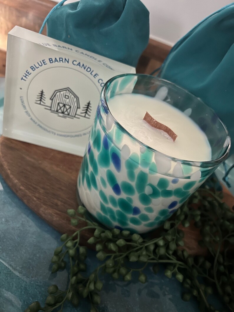 Puede incluir: Una vela de vidrio con un interior de cera blanca y una mecha de madera. El vidrio est&aacute; decorado con manchas azules y turquesas. Se ve una caja con el texto "THE BLUE BARN CANDLE CO.".