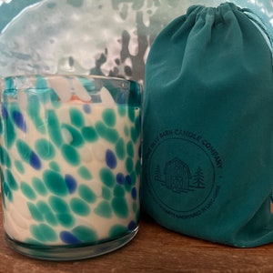 Puede incluir: Una vela de vidrio con un dise&ntilde;o moteado en tonos azules y turquesas, junto a una bolsa con cord&oacute;n turquesa. La vela est&aacute; sobre una superficie de madera, con un plato turquesa en el fondo. La bolsa tiene el texto "THE BLUE BARN CANDLE COMPANY".