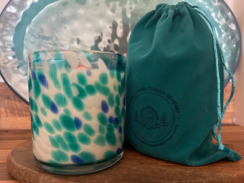 Puede incluir: Una vela de vidrio con un dise&ntilde;o moteado en tonos azules y turquesas, junto a una bolsa con cord&oacute;n turquesa. La vela est&aacute; sobre una superficie de madera, con un plato turquesa en el fondo. La bolsa tiene el texto "THE BLUE BARN CANDLE COMPANY".