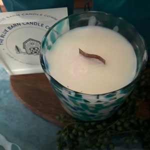 Puede incluir: Un portavelas de vidrio con una vela blanca y una mecha de madera. El portavelas tiene un dise&ntilde;o moteado azul y verde. Se ve una tarjeta con el texto "The Blue Barn Candle Company".
