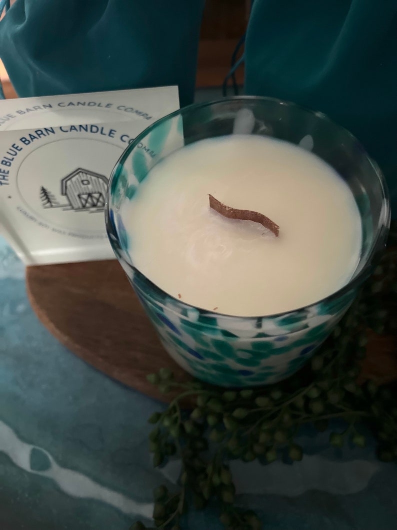 Puede incluir: Un portavelas de vidrio con una vela blanca y una mecha de madera. El portavelas tiene un dise&ntilde;o moteado azul y verde. Se ve una tarjeta con el texto "The Blue Barn Candle Company".