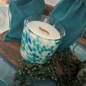 Puede incluir: Una vela de vidrio con un dise&ntilde;o moteado azul y verde, llena de cera blanca y una mecha de madera. La vela est&aacute; sobre una tabla de madera con una ramita de follaje verde. Dos bolsas turquesas con cord&oacute;n est&aacute;n en el fondo.