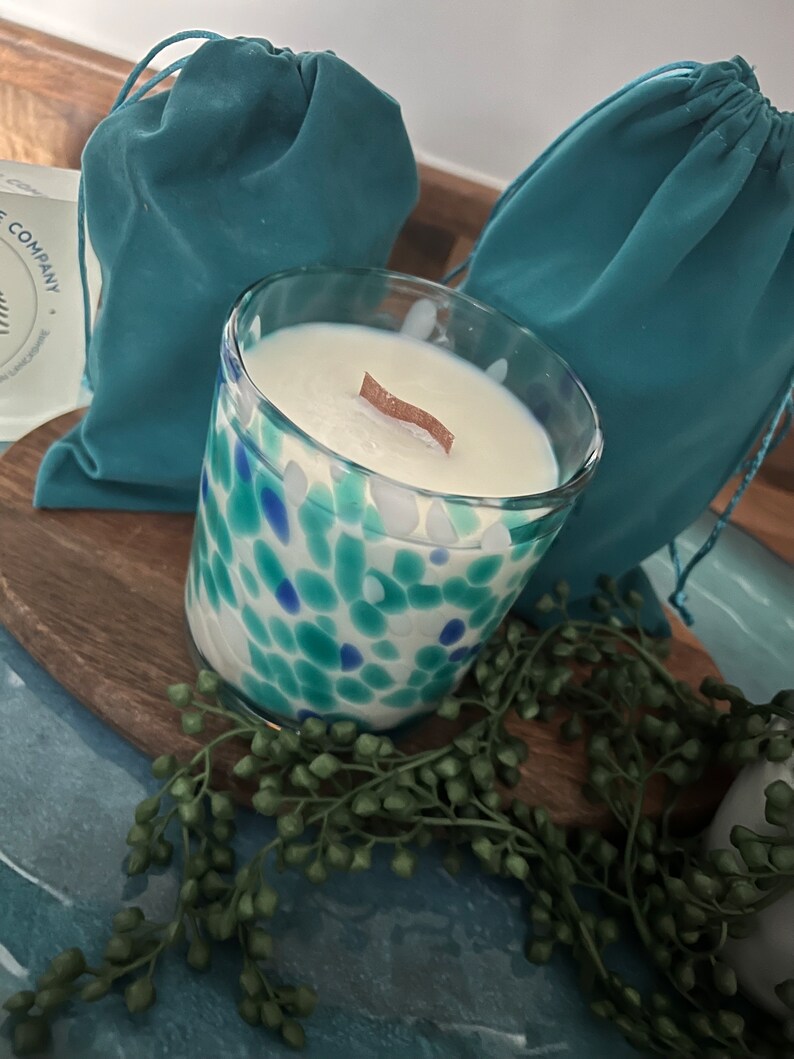 Puede incluir: Una vela de vidrio con un dise&ntilde;o moteado azul y verde, llena de cera blanca y una mecha de madera. La vela est&aacute; sobre una tabla de madera con una ramita de follaje verde. Dos bolsas turquesas con cord&oacute;n est&aacute;n en el fondo.