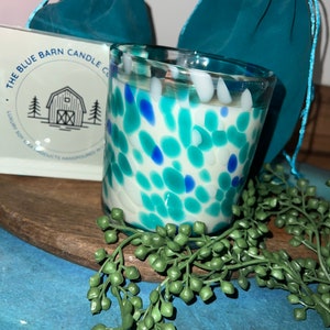 Puede incluir: Un portavelas de cristal moteado con detalles azules, turquesas y blancos. La vela est&aacute; rellena de cera blanca. El logotipo de The Blue Barn Candle Co. es visible en una caja blanca. Dos bolsas turquesas con cord&oacute;n est&aacute;n en el fondo.