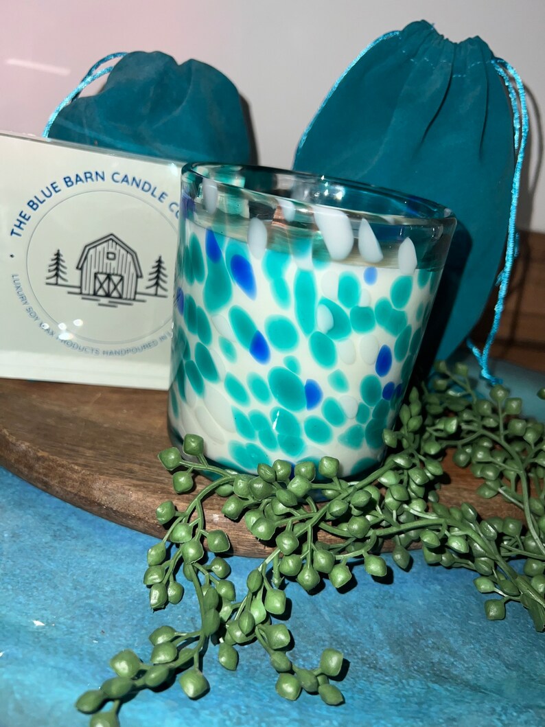Puede incluir: Un portavelas de cristal moteado con detalles azules, turquesas y blancos. La vela est&aacute; rellena de cera blanca. El logotipo de The Blue Barn Candle Co. es visible en una caja blanca. Dos bolsas turquesas con cord&oacute;n est&aacute;n en el fondo.