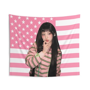 May include: A pink tapestry featuring a pink and white American flag design. A person with long black hair is wearing a pink striped shirt with green stripes. The person is touching their face with their finger.