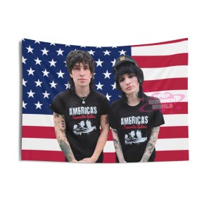 Puede incluir: Un tapiz con dos personas frente a una bandera estadounidense. Llevan camisetas negras con el texto "AMERICAS Favorite Ladies" y un gráfico. El fondo es la bandera de EE. UU. con rayas rojas y blancas y un campo azul de estrellas.