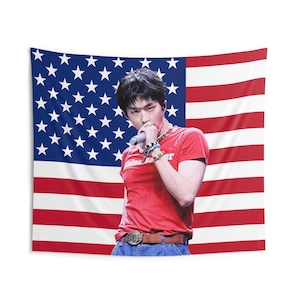 Puede incluir: Una persona con una camiseta roja con el texto "Lost Lover" está de pie frente a un tapiz de la bandera estadounidense. La persona sostiene un micrófono. El tapiz muestra las estrellas y las rayas de la bandera estadounidense.