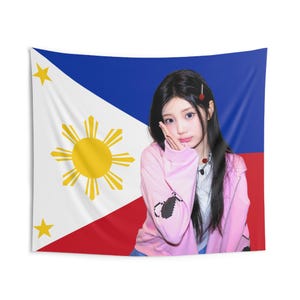 May include: A tapestry featuring the flag of the Philippines with a red and blue background and a yellow sun and stars. A person wearing a pink jacket and white shirt is in front of the flag.