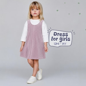 May include: A young girl wearing a white long-sleeve shirt under a pink corduroy jumper dress. The dress has vertical stripes. The image includes the text "Dress for girls 12M-14Y". She is wearing white sneakers.