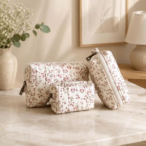 Könnte beinhalten: Ein Set aus drei gesteppten Kosmetiktaschen in verschiedenen Größen. Die Taschen sind weiß mit einem sich wiederholenden Muster aus kleinen roten Blumen. Jede Tasche hat einen Reißverschluss und einen dunkelbraunen Reißverschluss.