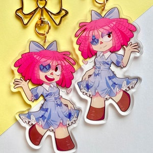 Puede incluir: Dos llaveros de acr&iacute;lico transparente con un dise&ntilde;o de mu&ntilde;eca de dibujos animados. La mu&ntilde;eca tiene pelo rosa, un vestido azul y botas marrones. Los llaveros est&aacute;n sujetos a cierres dorados en forma de lazo. El fondo es una mezcla de amarillo y blanco.